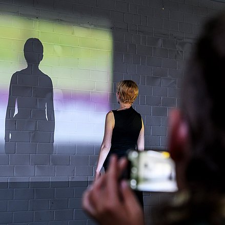 Eine Person steht vor einer Wand und wirft durch buntes Licht Schatten an die Wand