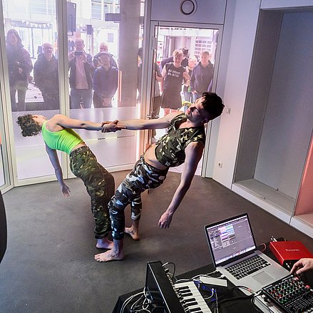 Zwei Personen tanzen vor einem DJ mit Mischpulten und Zuschauer schauen durch eine Fensterscheibe zu