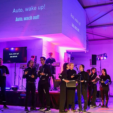 Ein Chor singt unter bunter Bühnenbeleuchtung den Text „Auto, wake up!“ auf Englisch und Deutsch, der groß projiziert wird.